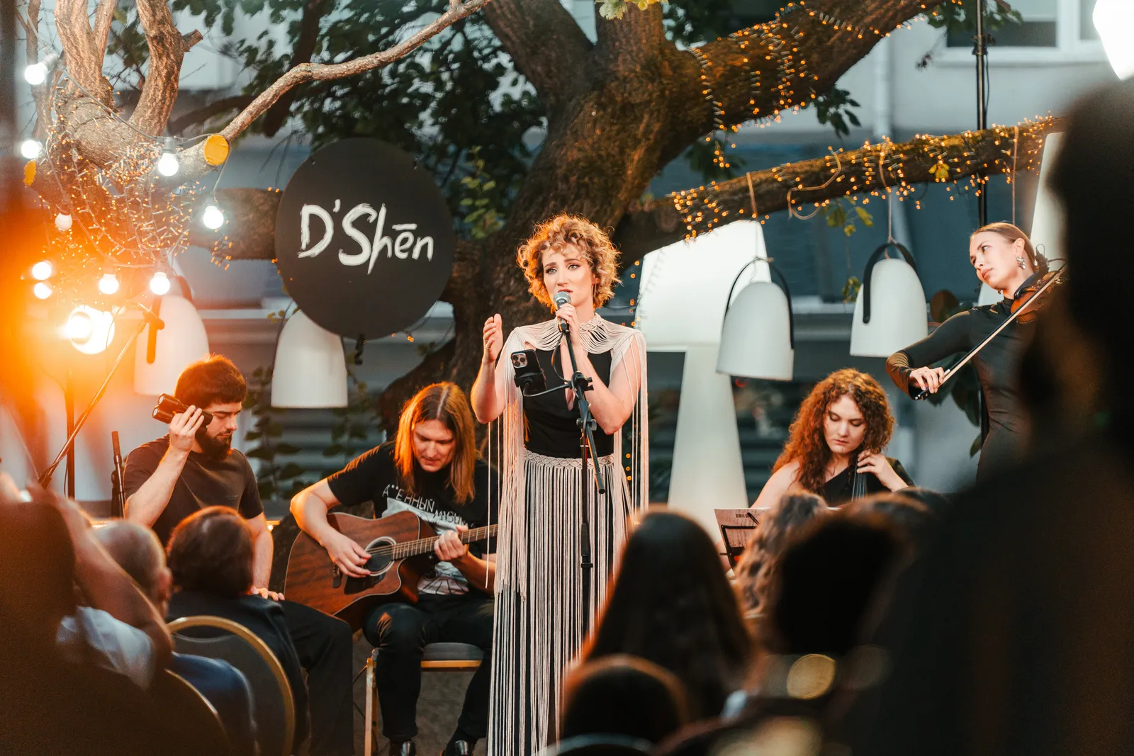 The full band under a tree strung with fairy lights, crowd silhouettes in the foreground